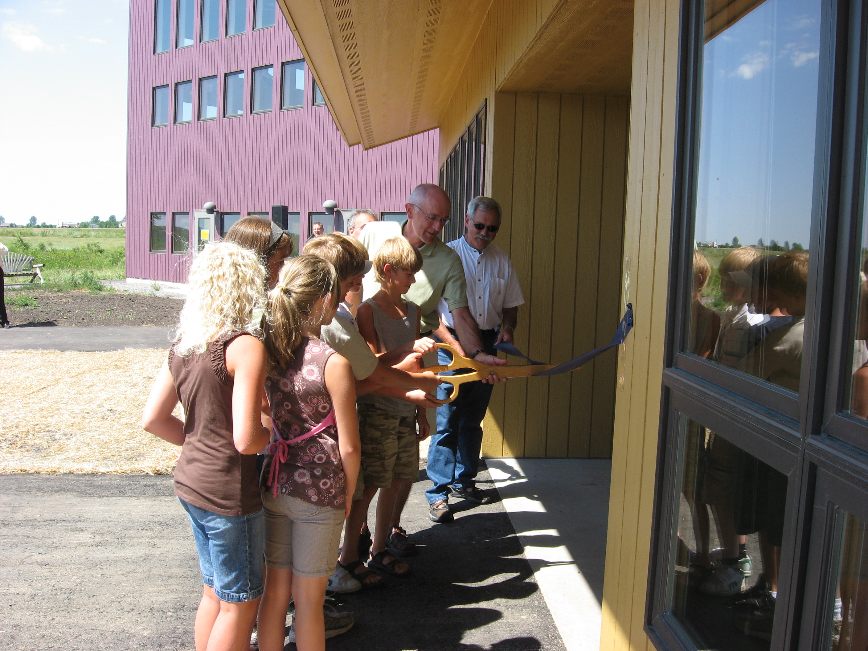 Adults and students use a large wooden scissors to cut a ribbon outside a new classroom
