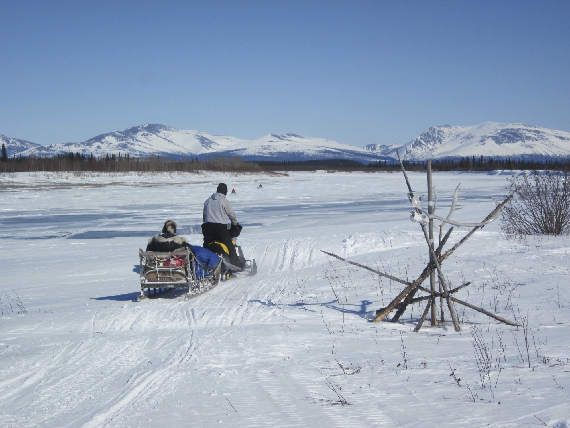 A person drives a snowmobile over past a wooden trail stake, towing a wooden sled with a passenger sitting inside