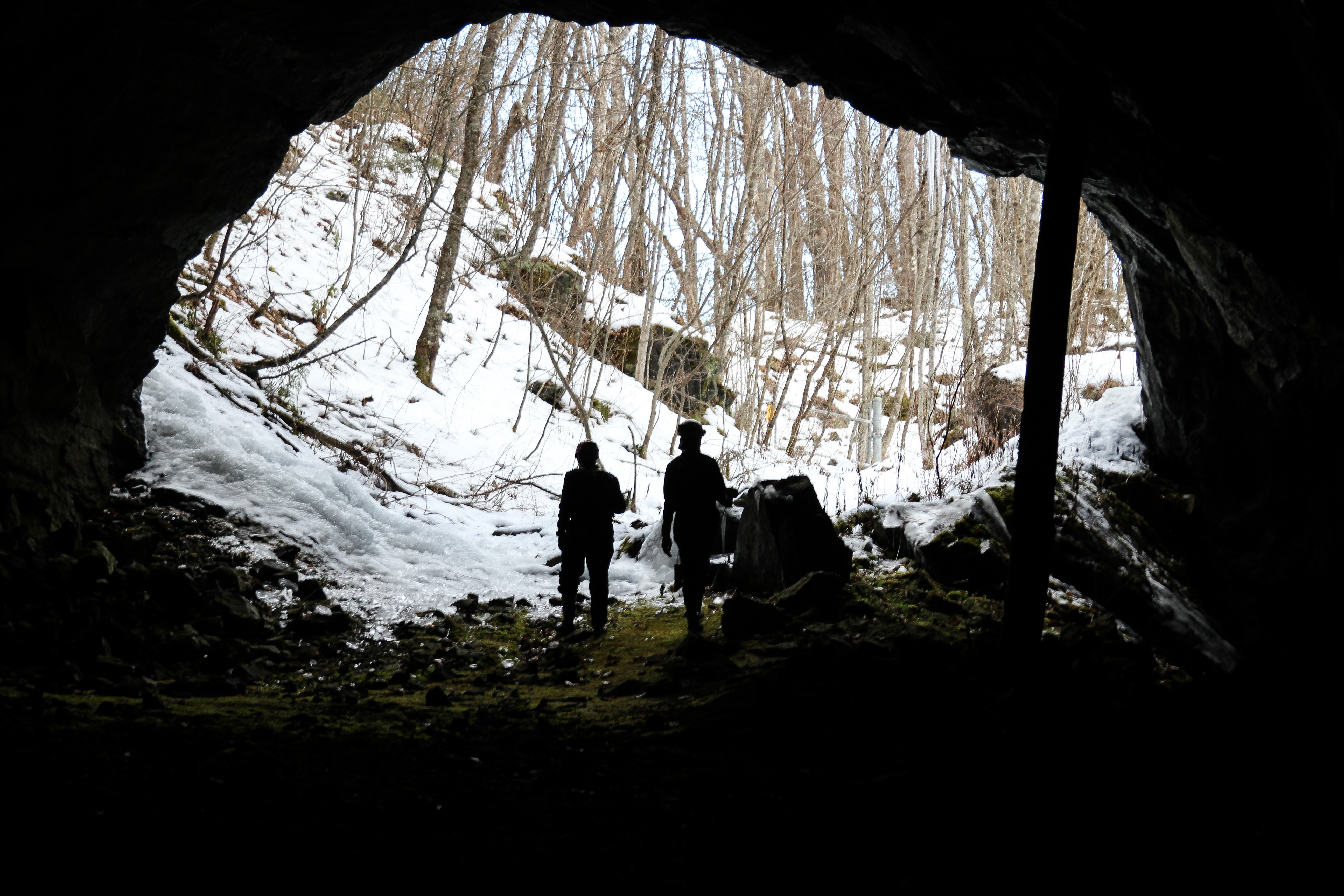Two people exiting through the mouth of a mine, entering a snow-covered forest