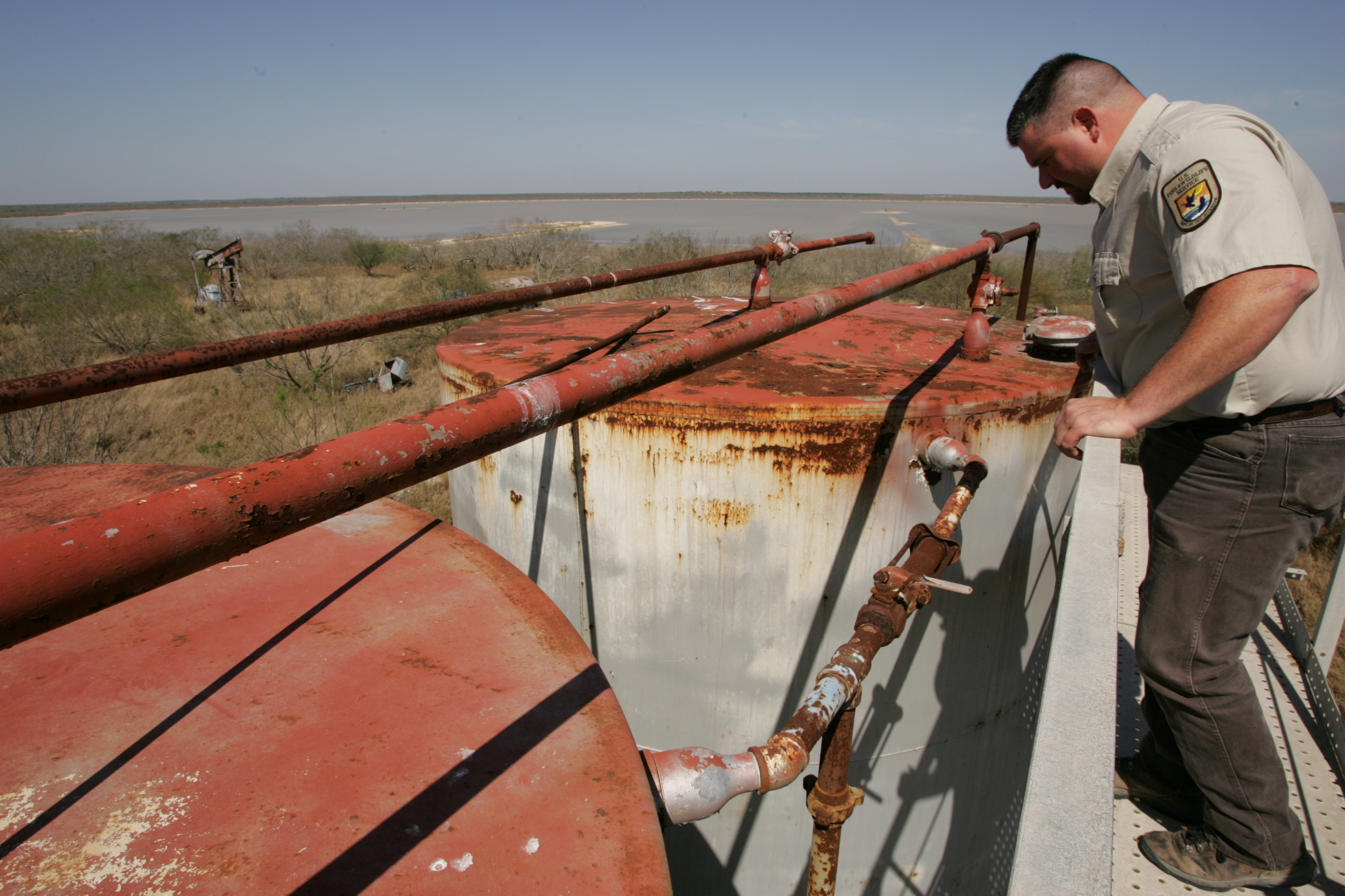 A man in a Fish and Wildlife Service uniform looks at rusting oil tanks that sit on land.