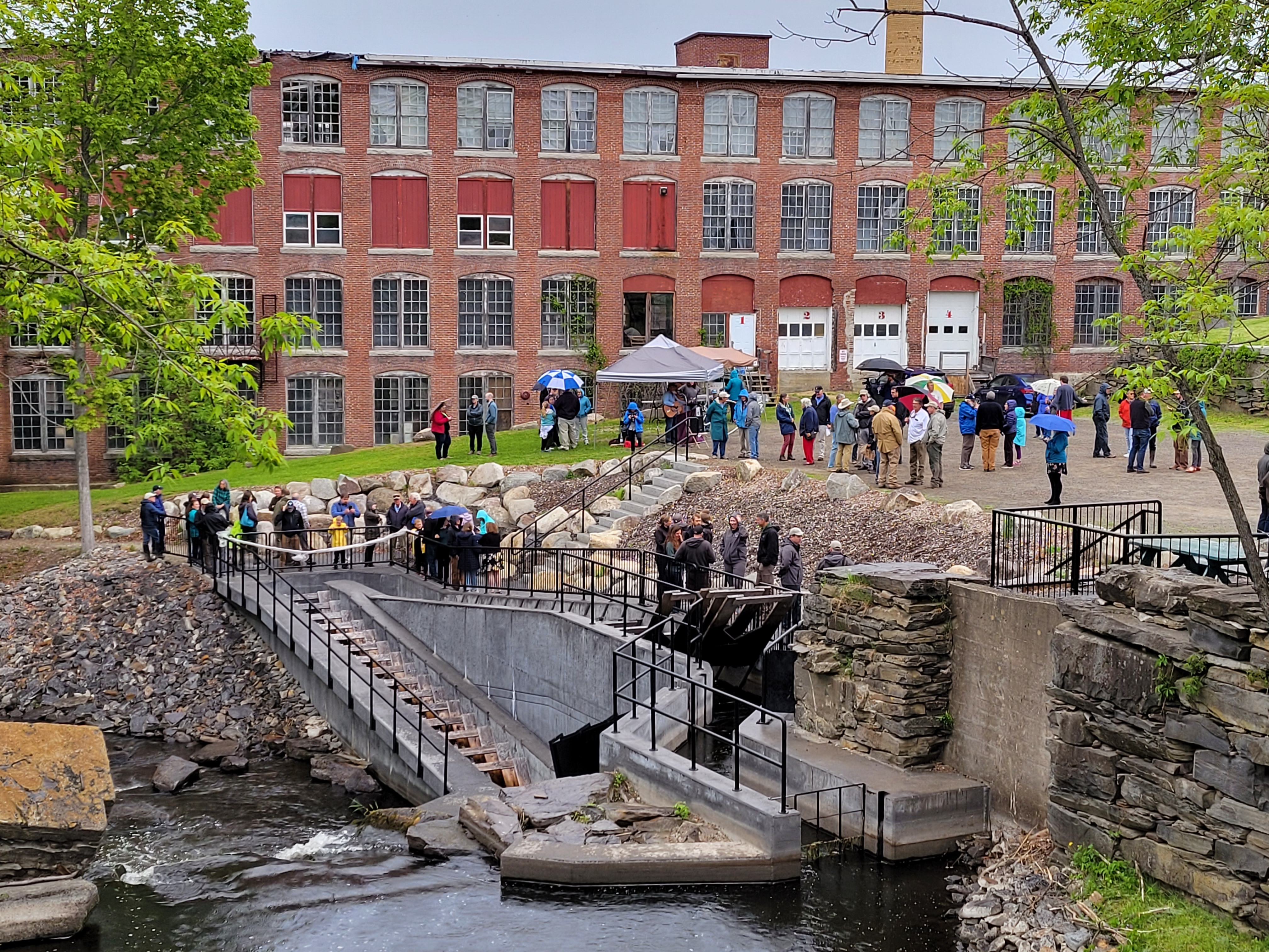 People gather around a fishway structure before a large, old mill building
