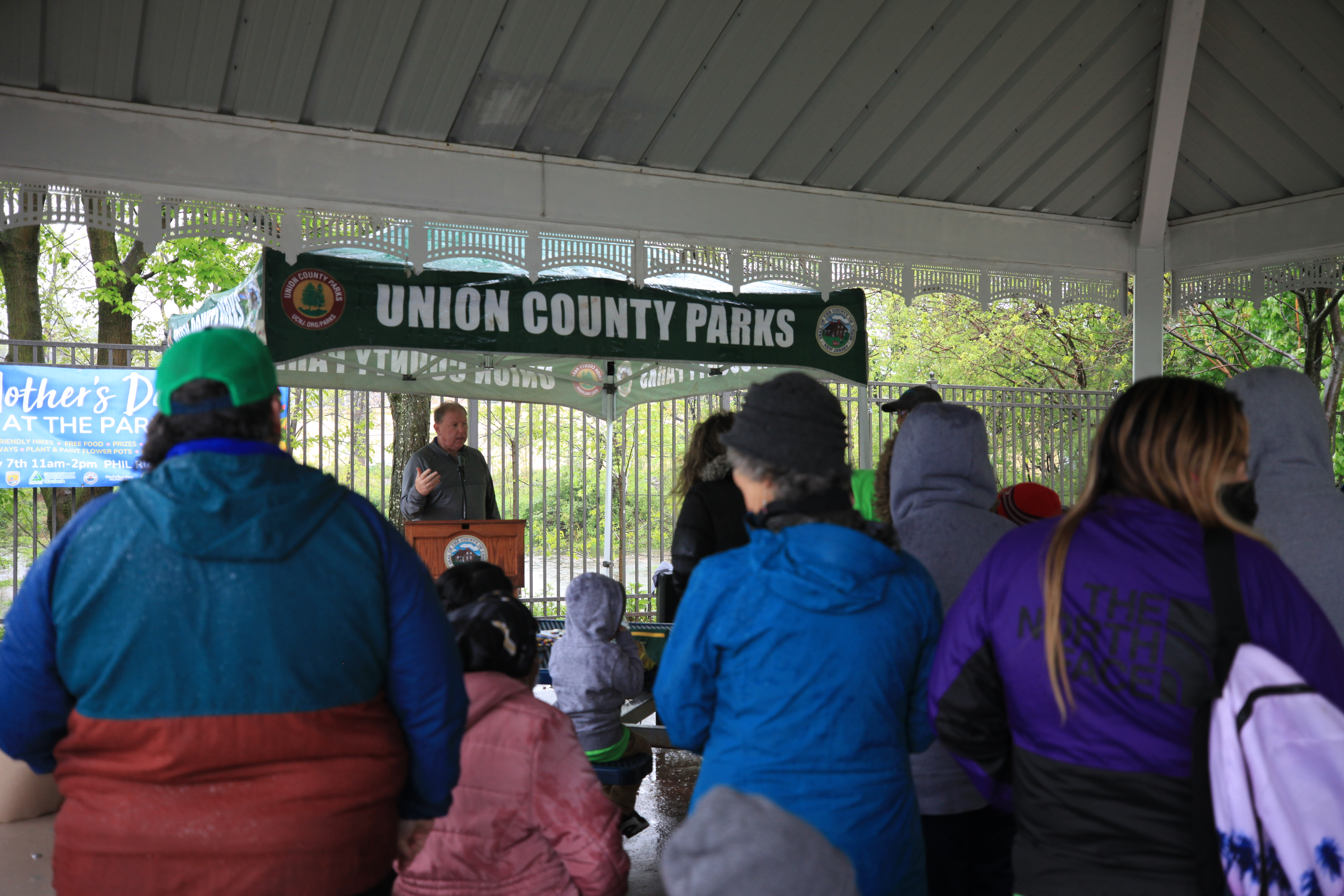 a man speaks at a podium in front of a crowd of people. A tent banner above him reads "UNION COUNTY PARKS"