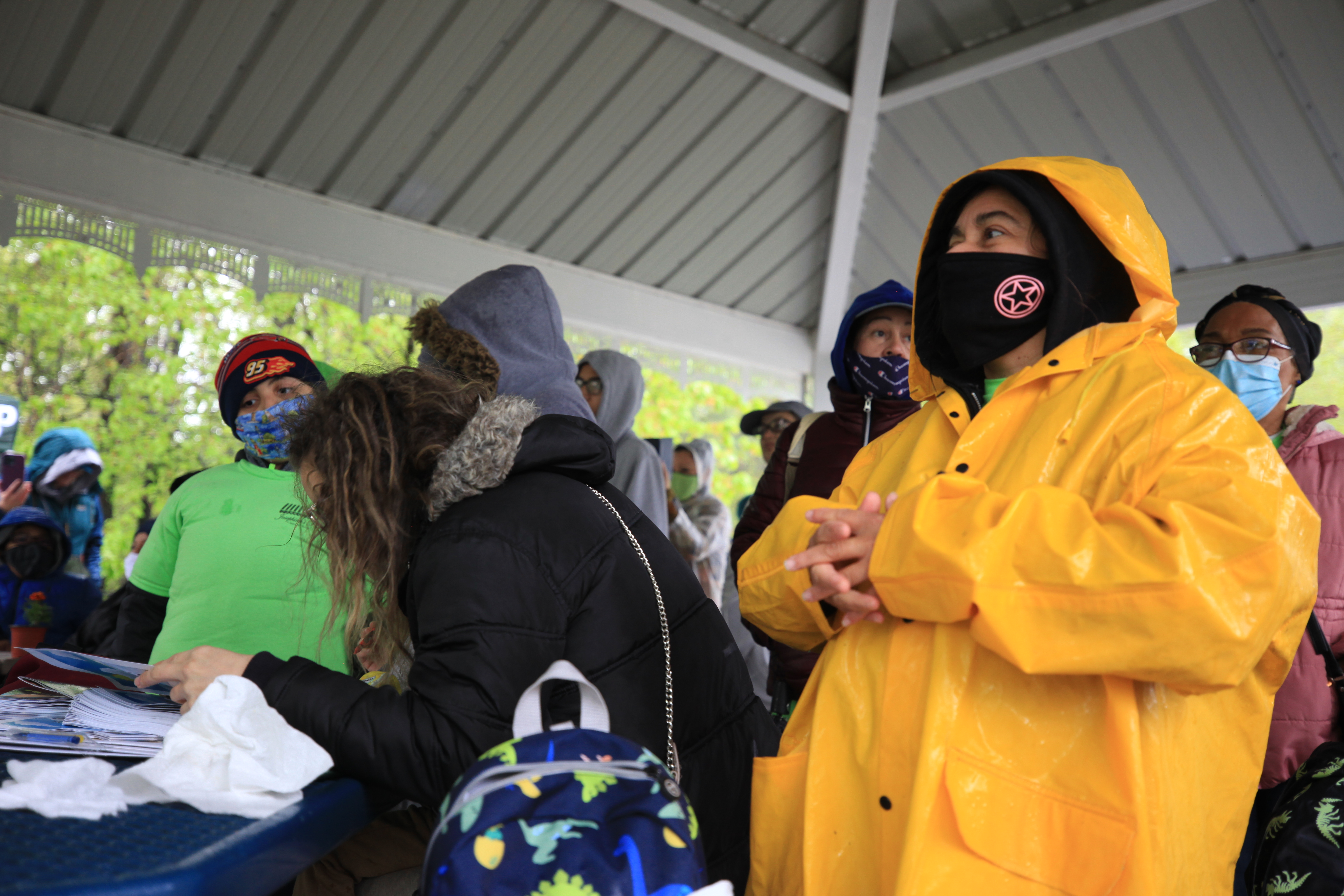 people stand under a pavilion in rain jackets and masks.
