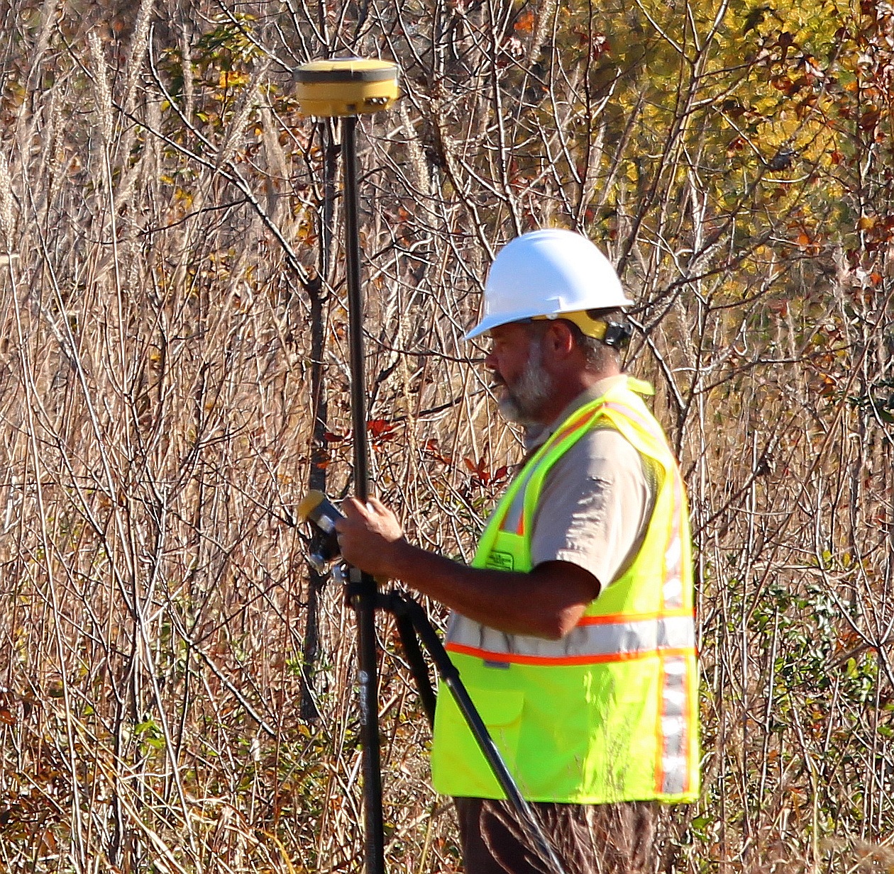 A wearing a bright yellow safety vest and a white safety helmet holds a surveying tool in brush