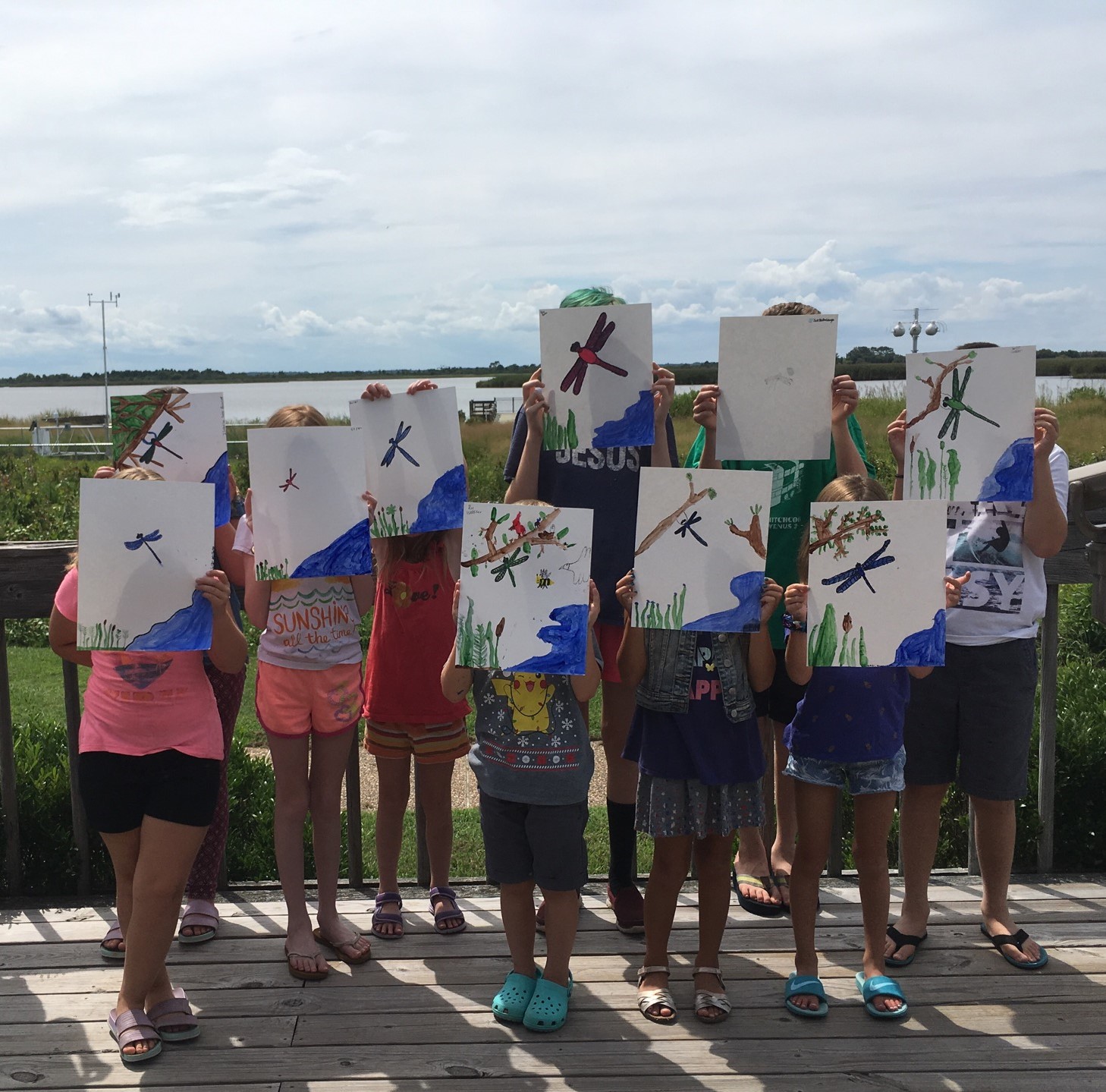 Ten children hold paintings of dragonflies in front of their faces. The children are on a deck with marsh and water in the background.