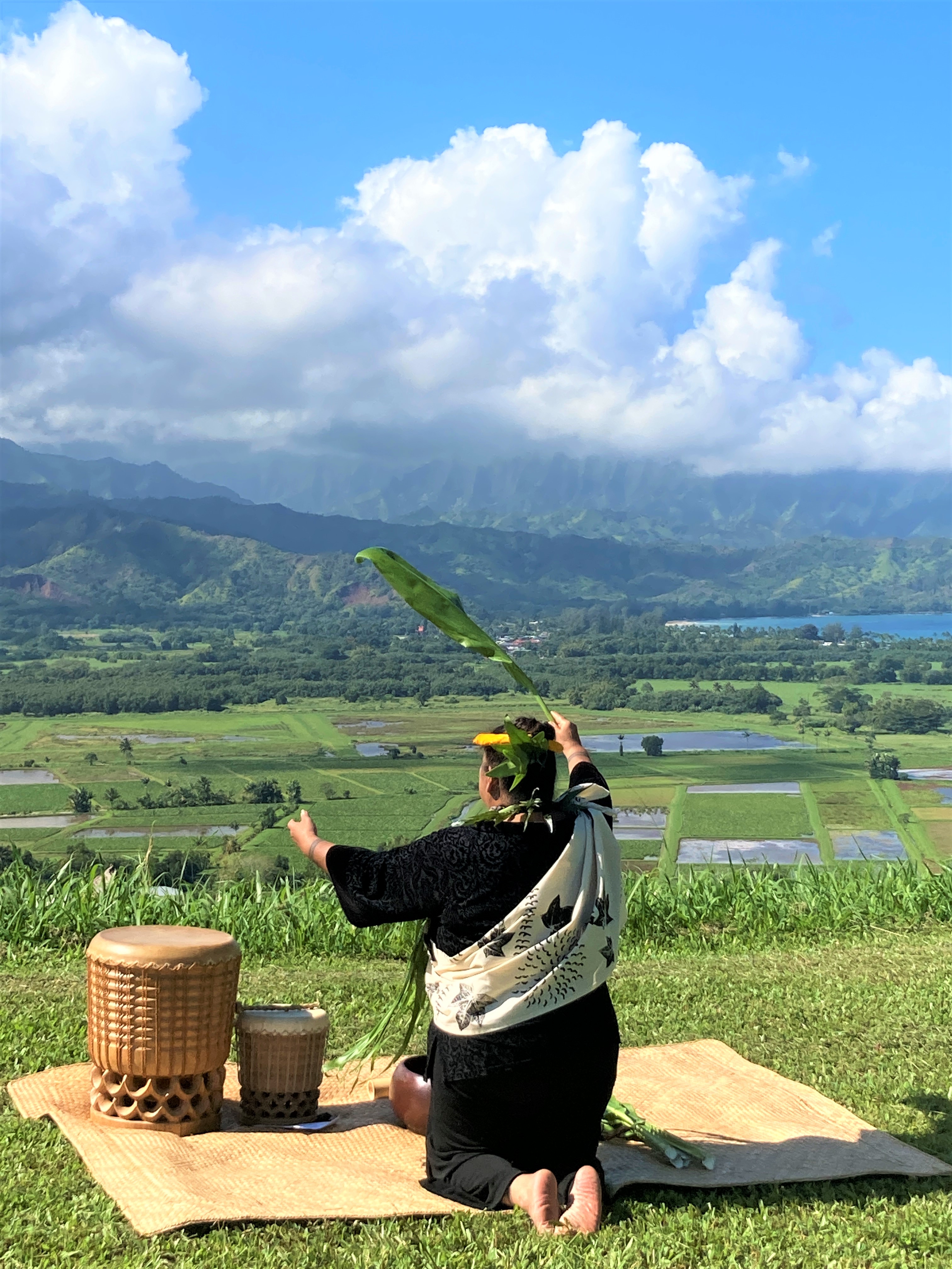 A woman kneels on the ground. She is sitting on a lush grassy hill that overlooks taro fields, mountains, and the ocean.
