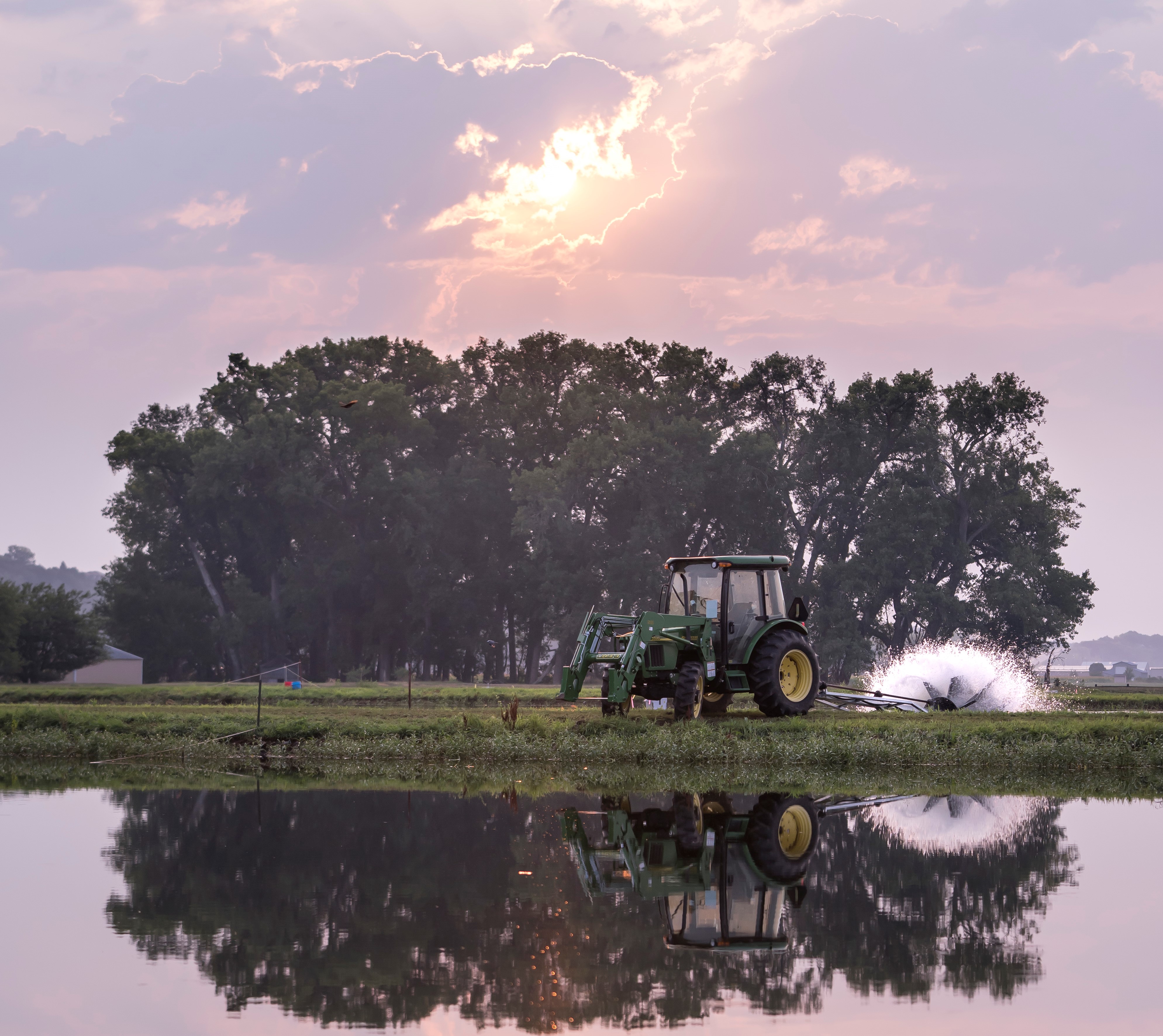 A large green tractor pulls a piece equipment to aerate a shallow pond with the sun peaking through the morning clouds and a stand of trees in the distance