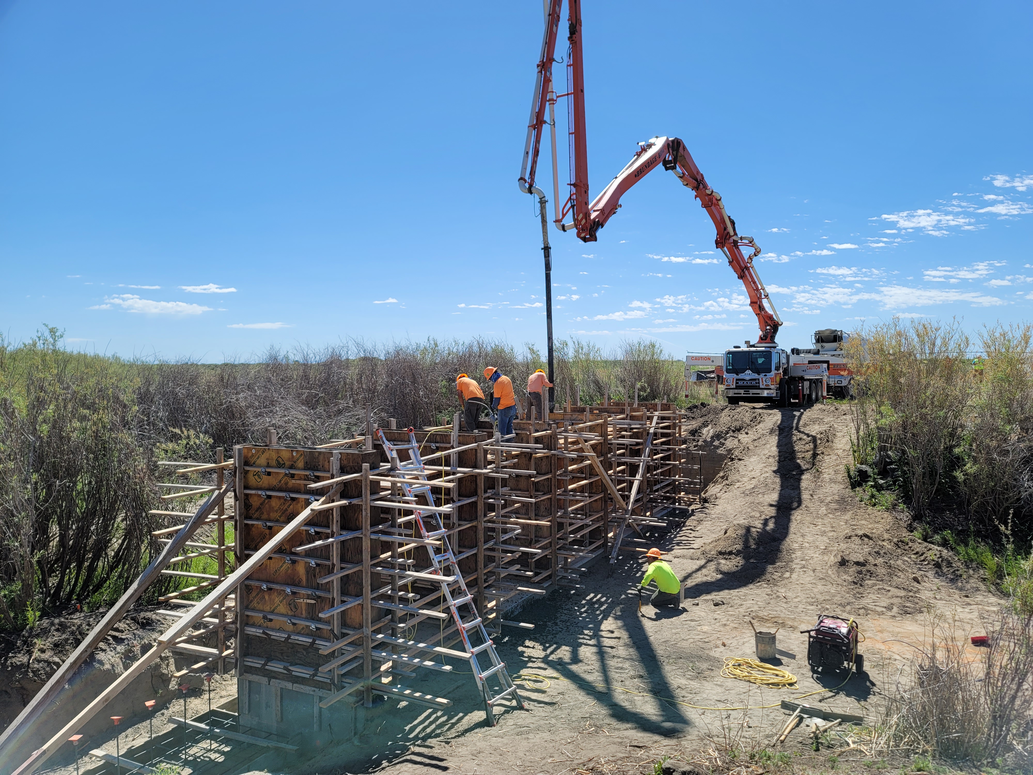 Construction workers build a structure at an Idaho refuge, with a truck using a crane arm to pipe in materials, on a clear day.