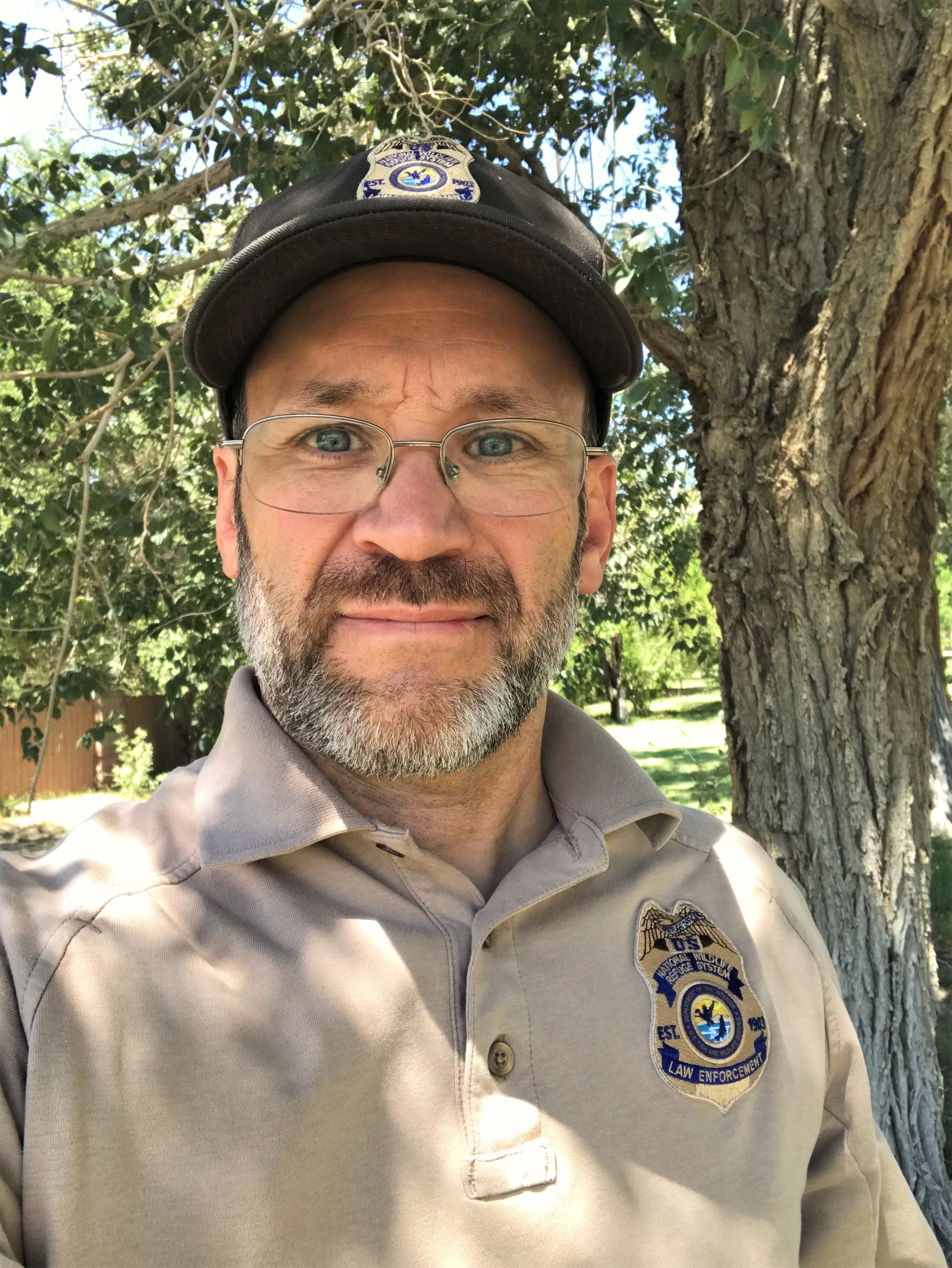 A smiling man with a beard and glasses in a U.S. Fish and Wildlife Service cap and uniform standing near a tree