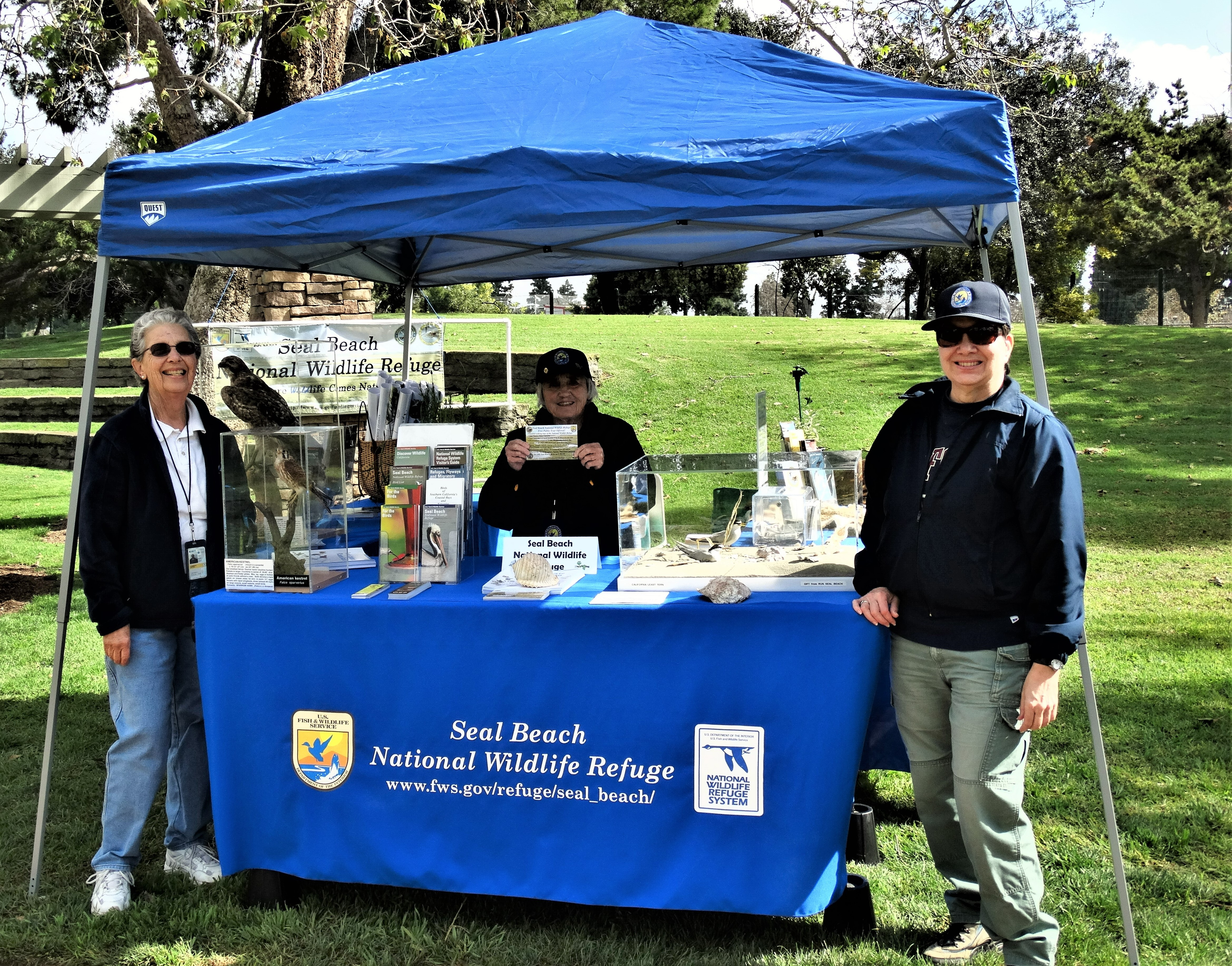 Three women stand at a blue-tented booth ready to engage visitors. The blue bunting on the booth has the words "Seal Beach National Wildlife Refuge" on it.