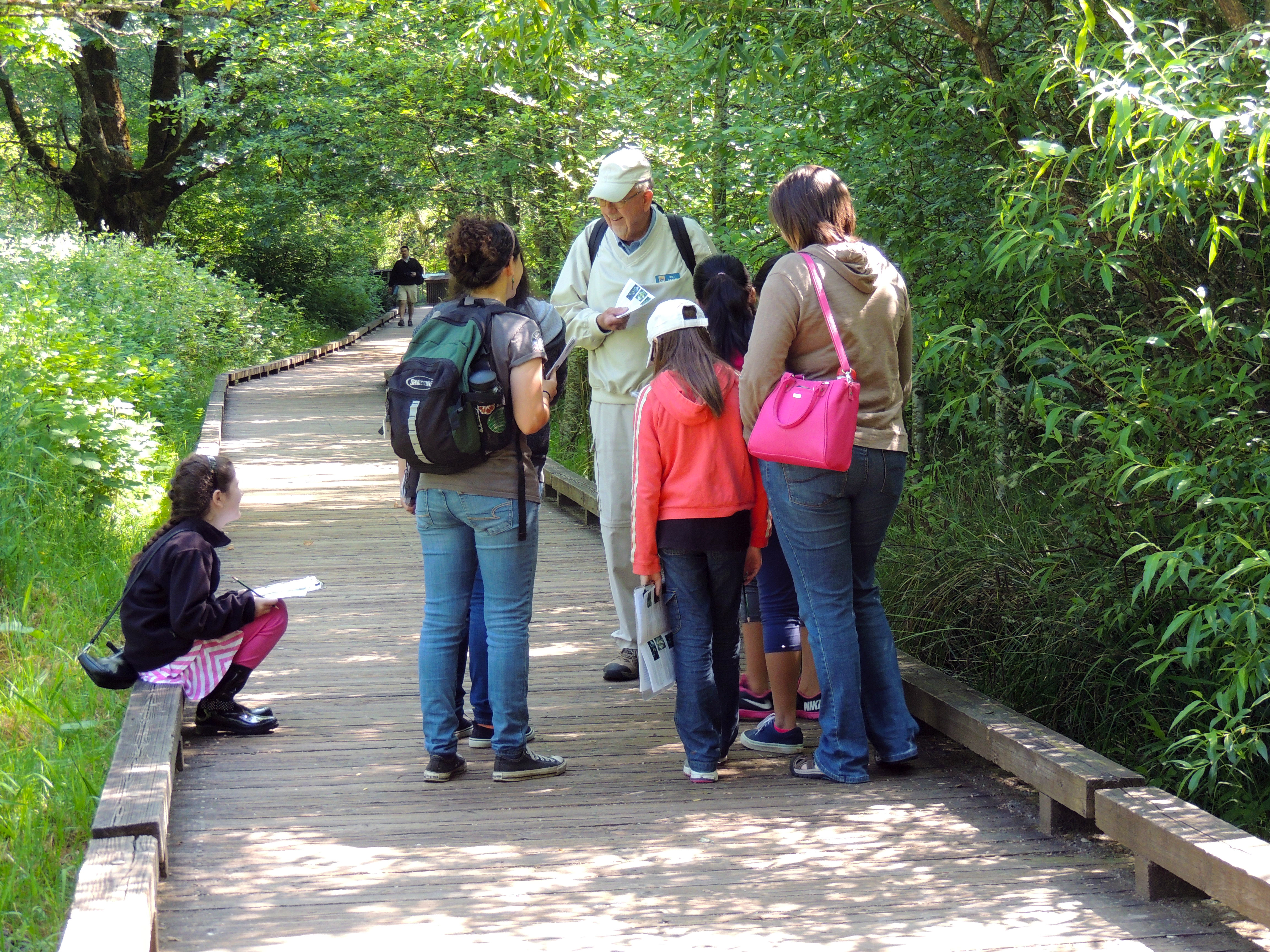 An older man in a white cap with a nametag smiles while talking with a small group of youth and another adult on a boardwalk trail in a wooded area.