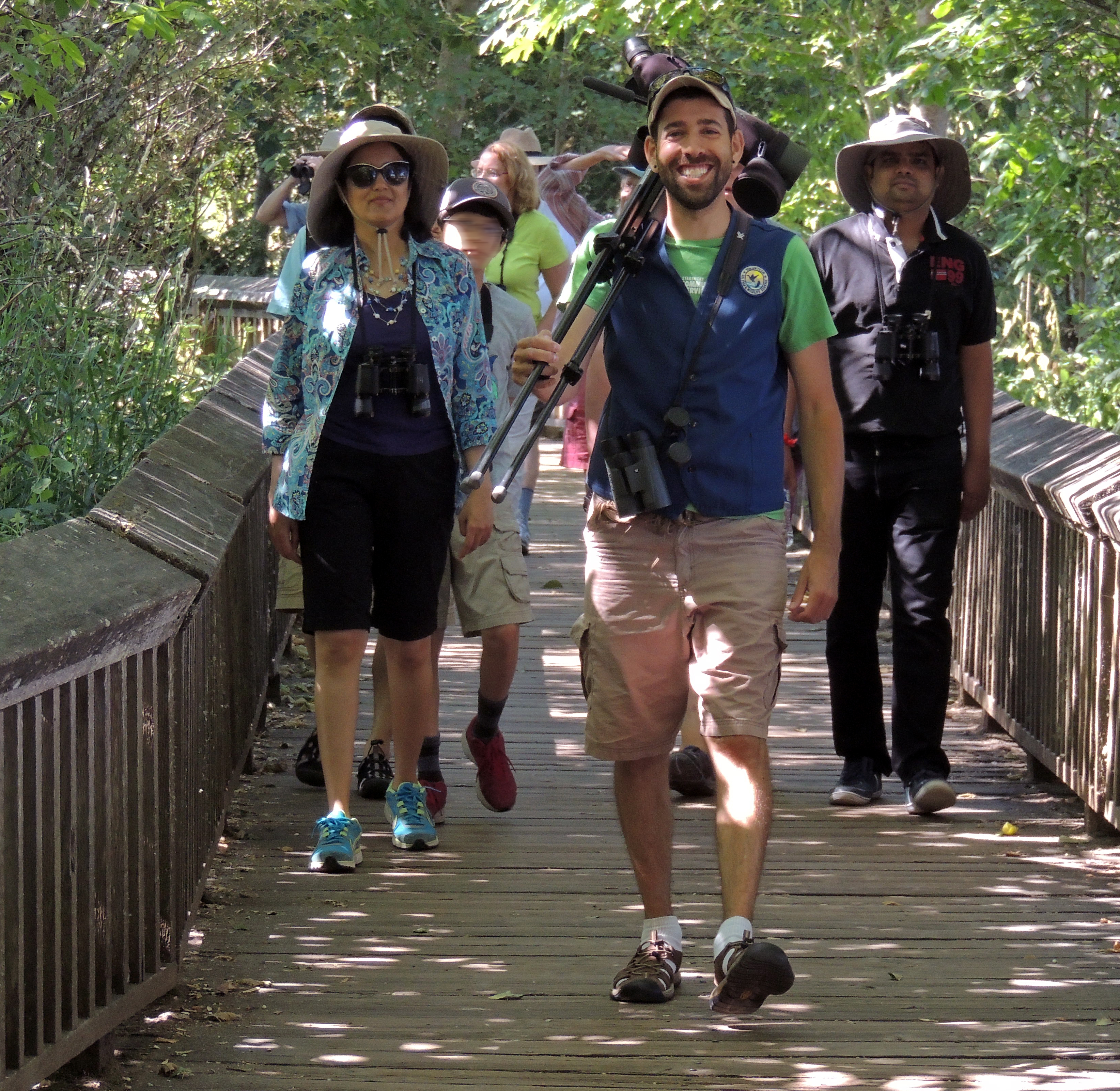 A grinning young man in a volunteer vest and shorts with a scope over his shoulder leads a group of visitors in summer wear along a boardwalk trail.