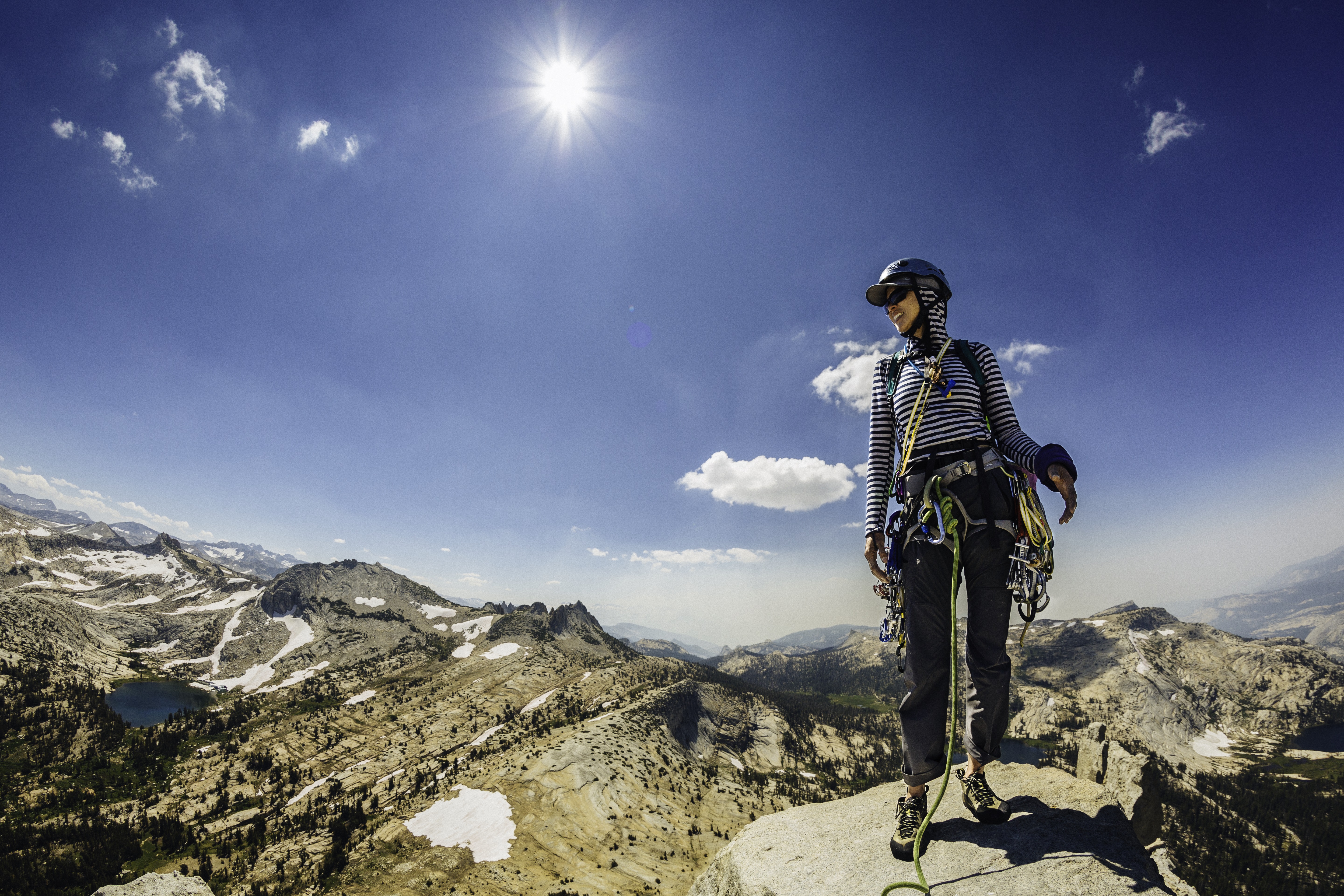 A woman in climbing gear and a helmet looks down at a park covered in pine trees from the summit of a mountain