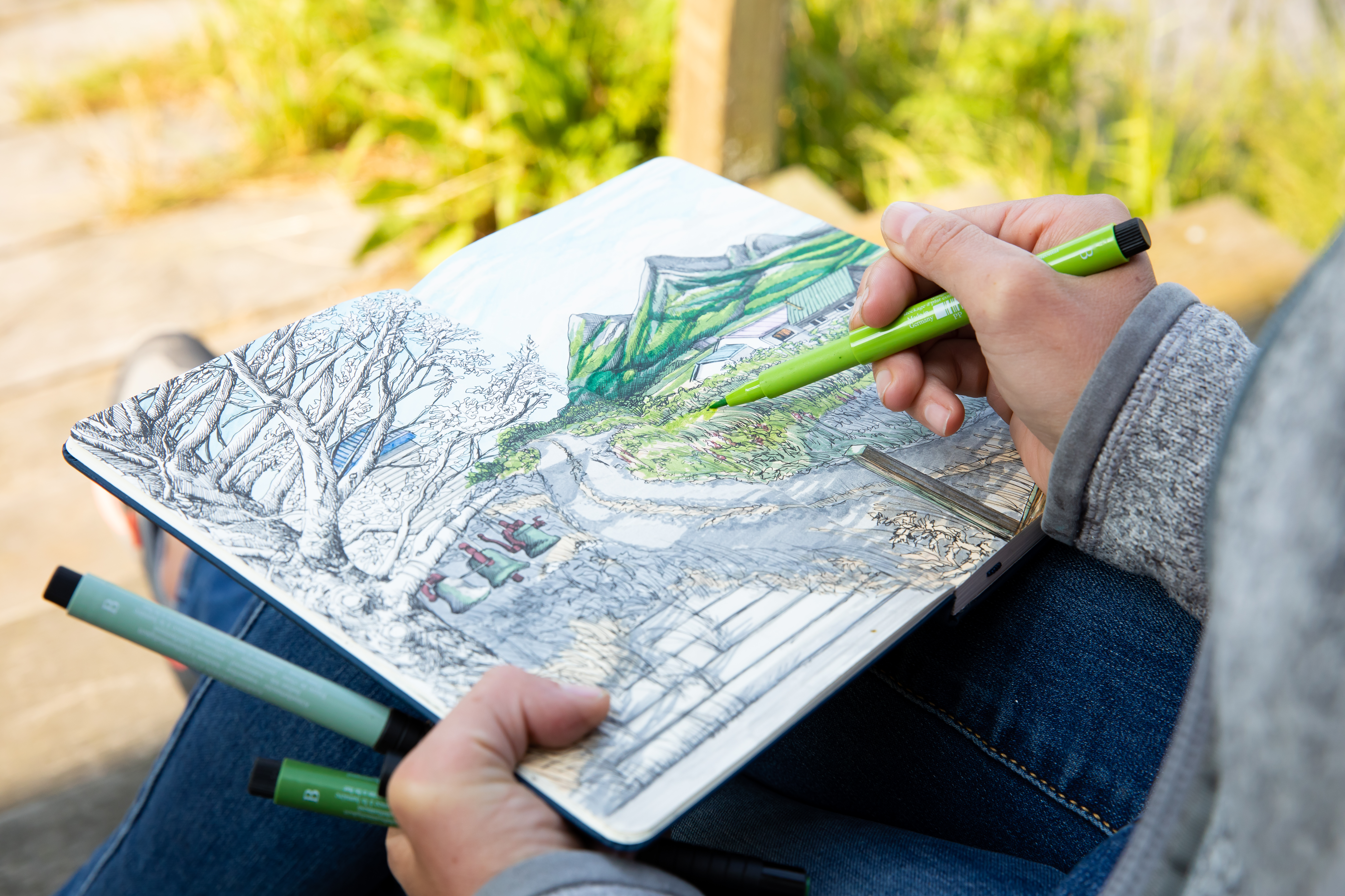 A hand holding a green felt tipped pen coloring in an illustration in a sketchbook of a mountainous scene
