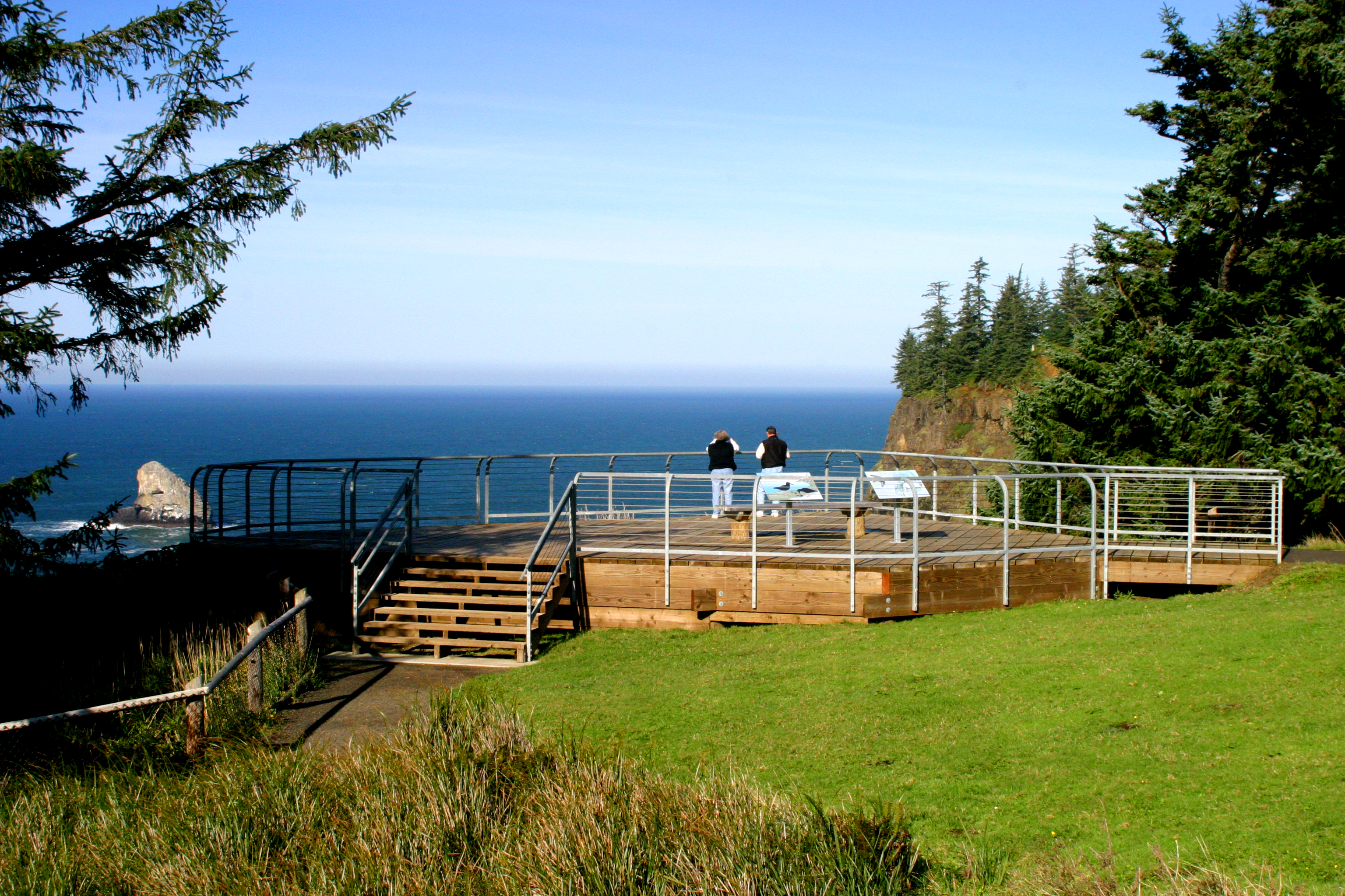 Two Visitors stand on a wooden deck overlooking sheer cliffs, rocky sea stacks, and the roiling Pacific.