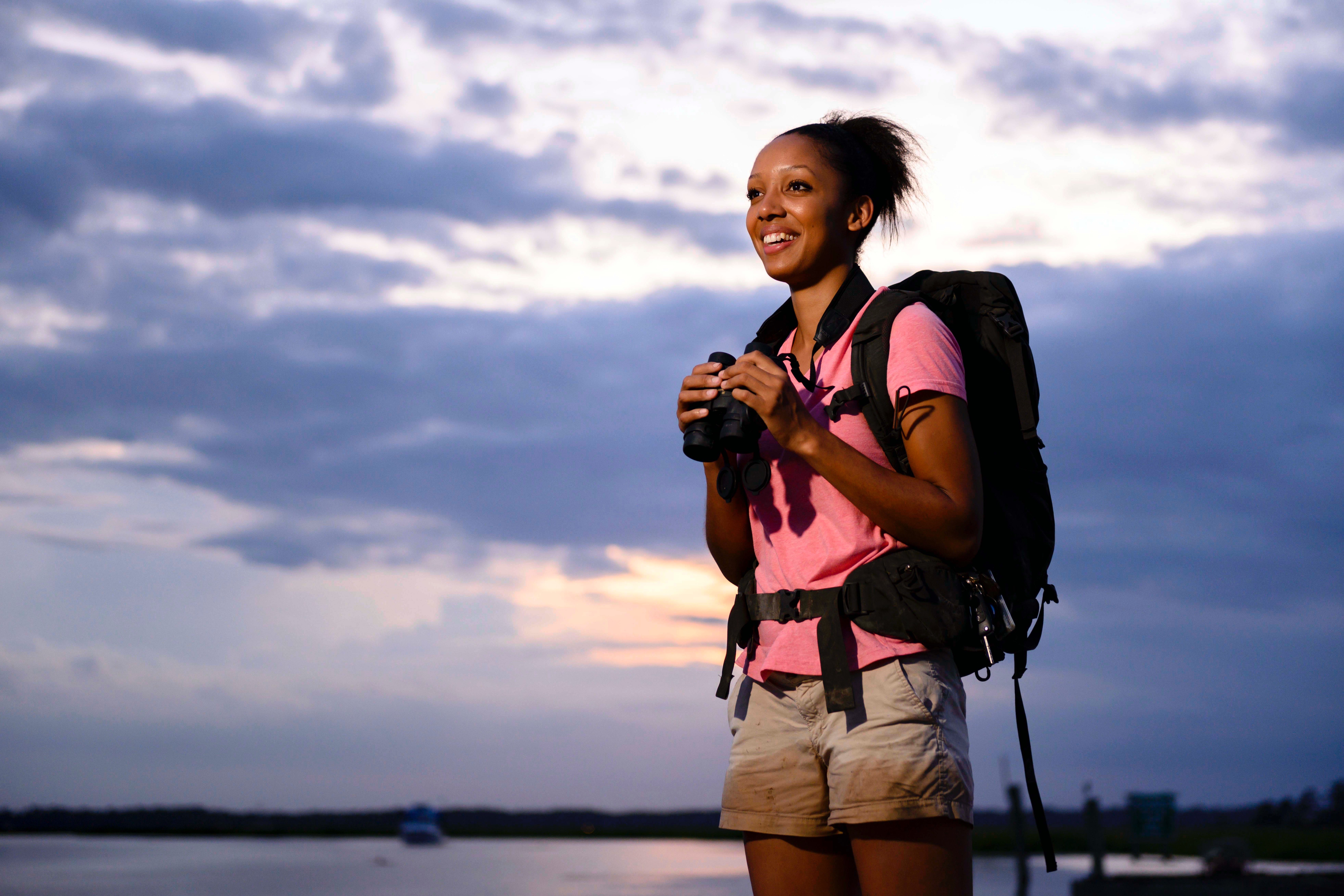A young Black woman wearing a backpack and holding binoculars and smiling, looks off into the distance under a cloudy blue sky.