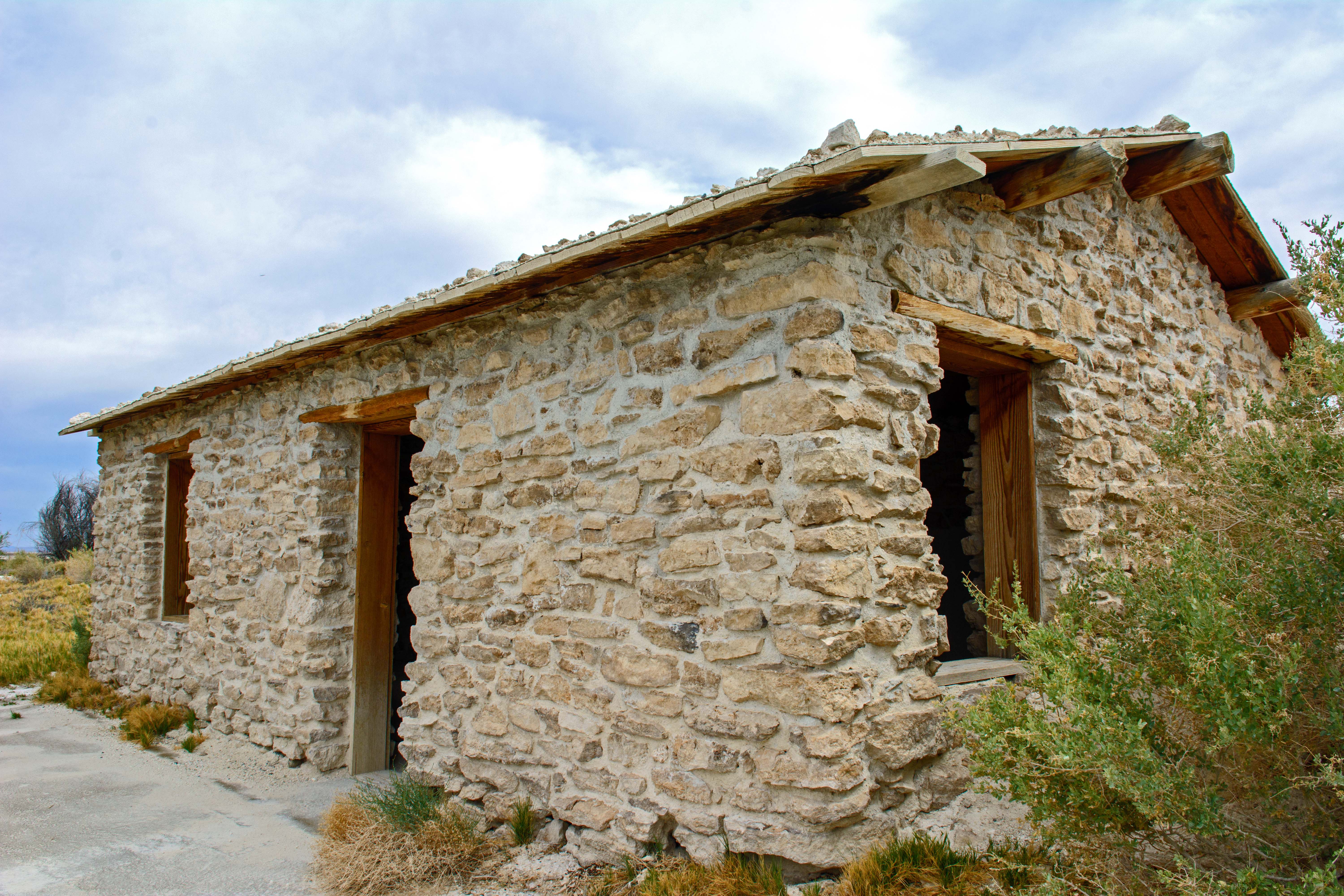  a white stone cabin with a door and two windows. Saltbush growing on right side of cabin