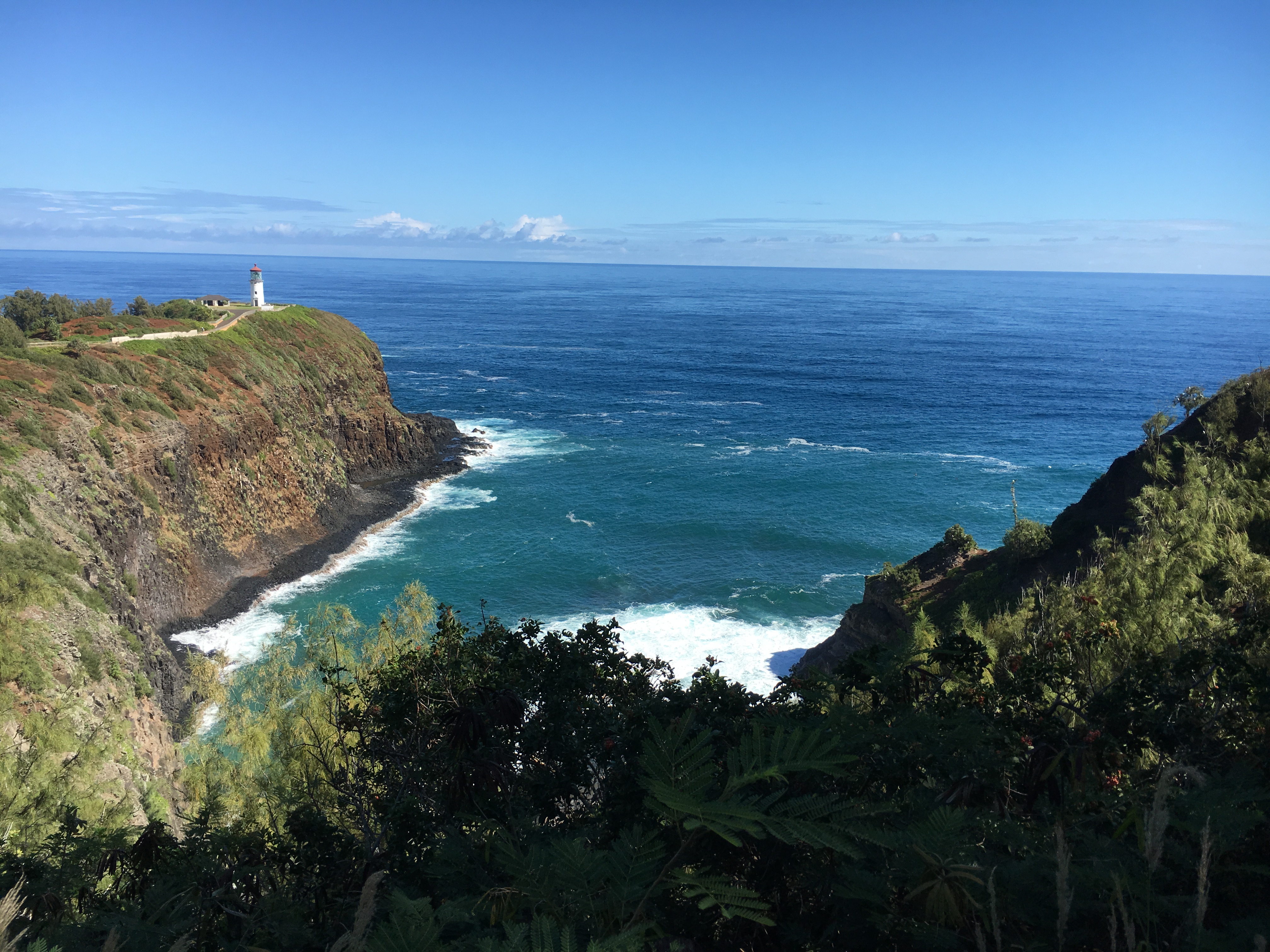 Kiluea Point National Wildlife Refuge lighthouse sits on a point. The photo shows a narrow inlet where the blue waters of the Pacific meet the high cliff faces of the refuge. Blue sky gleams in the back with contrast against the bright green of the forest. 