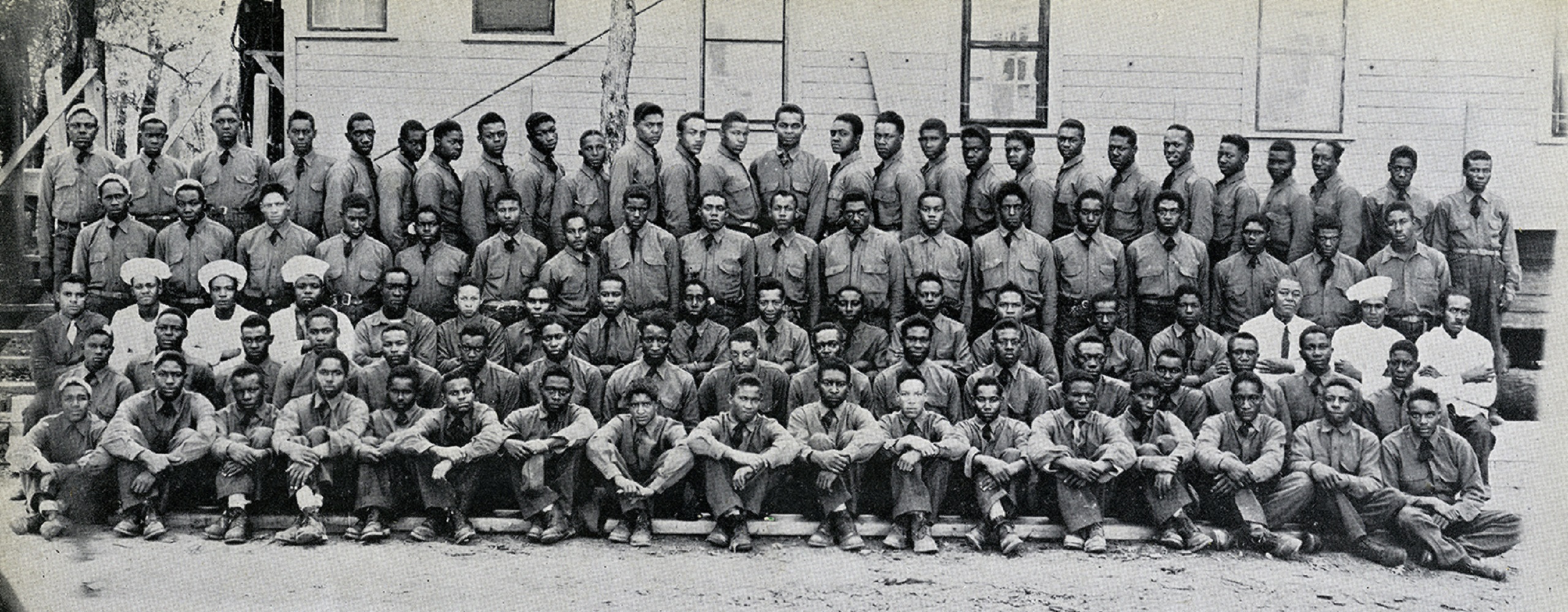 Black and white photo of more than 100 young African American men in uniform posing in front of a building