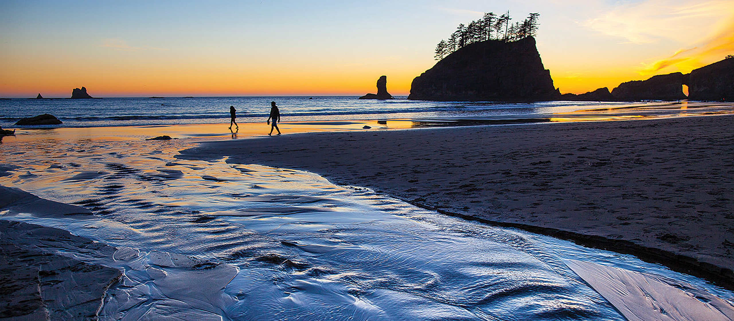 A stream flow down a beach to the ocean with silhouettes of humans walking and a distant sea stack at sunset