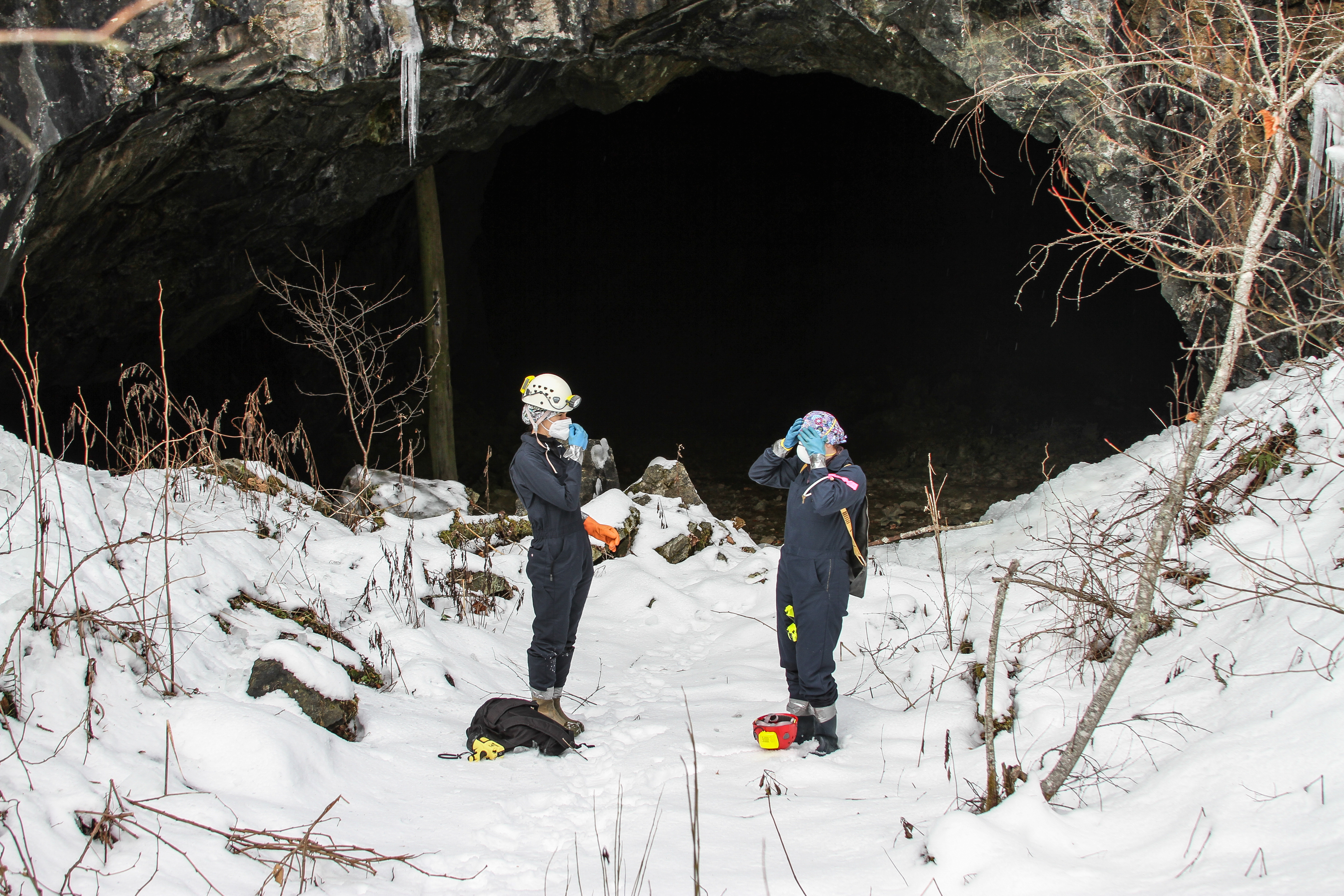Two people stand in the snow outside a mine entrance, making adjustments to their helmet and face mask