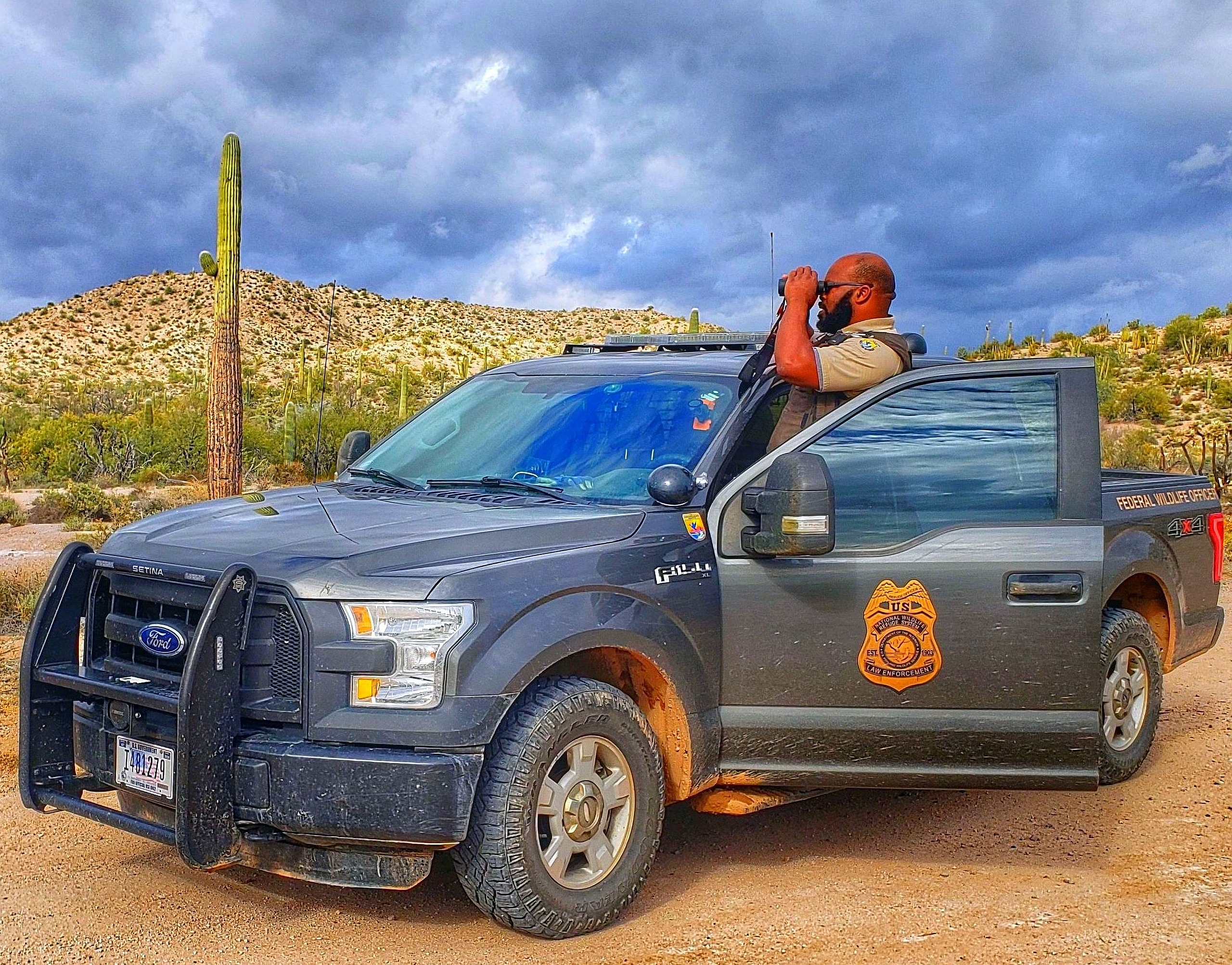A law enforcement officer standing in his truck in a desert setting looking through binoculars