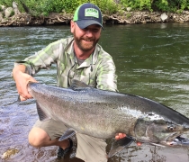 A smiling man in shorts and ballcap kneels at the edge of a river with a huge summer chinook salmon held out to the camera.