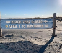 A somewhat see through banner is being held up by two poles. The banner is on a beach and reads "Beach Area Closed April 1 to September 30". 