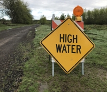 Bright yellow sign says "high water" to indicate high water levels on trail; sign is posted low to the ground next to gravel trail