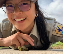 A woman in a U.S. Fish and Wildlife Service uniform smiling as she lies on the ground with a tiny turtle crawling in front of her