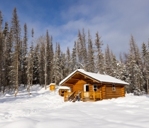 A wood cabin sands in the snow at the edge of a forest.