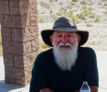 An older man with long white beard wearing a hat and dark blue shirt posing with a camera. 