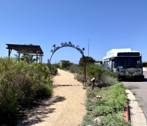 A trail that leads to the native pollinator garden as written on an overarching metal arch above the trail. Next to this is a shuttle bus. 