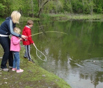 An adult helps a young girl reel in a fish while another child helps net the fish. They are standing on the edge of the water in a green natural setting.
