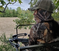 View of a field looking over the left shoulder of a hunter dressed in camouflage with a crossbow in a tree stand.