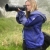 A woman in a purple rain coat holds a large camera looking through the lens. She is in a grassy area and the air is heavy with fog. Some trees can be seen in the fog behind her. 