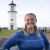 A blond woman in her 20's in a blue uniform stands in front of a lighthouse. She is smiling. 