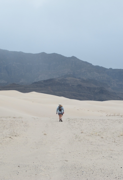 a woman walks across white sand dunes headed toward rugged mountains