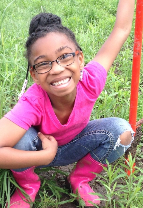 A smiling girl looks up from the grassy area where she is planting milkweed.
