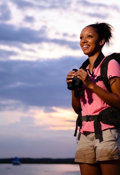 A young Black woman wearing a backpack and holding binoculars and smiling, looks off into the distance under a cloudy blue sky.