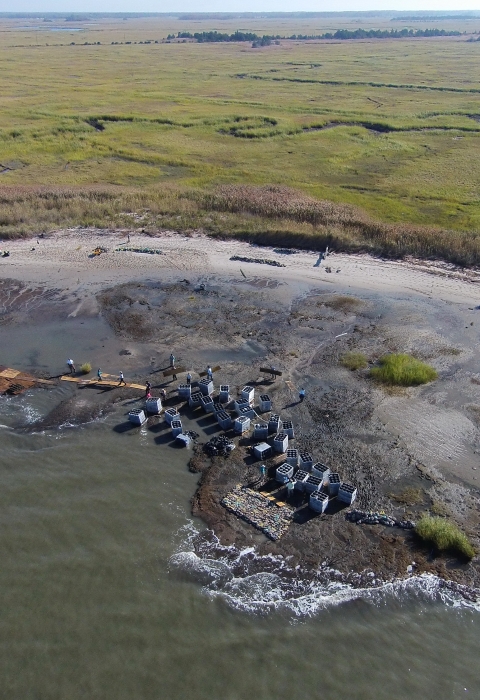 In this aerial image, Gandys Beach is visible in the foreground, as are a handful of volunteers and project staff and large square, gray containers filled with reef materials. A green marsh is visible behind the beach.