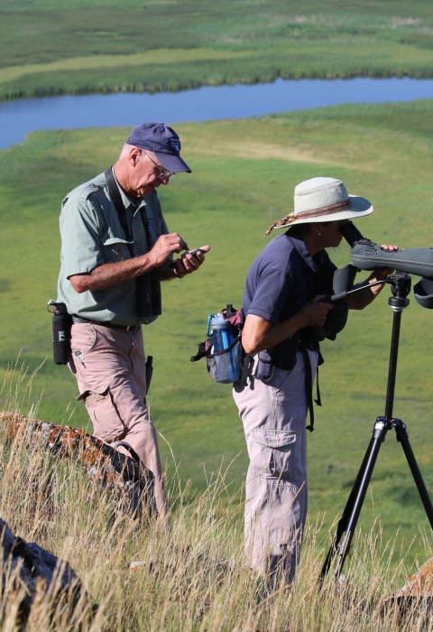 Volunteers Chuck and Betty Mulcahy, wearing hats and hiking clothes, survey trumpeter swans and cygnets weekly from atop a butte at National Elk Refuge in Wyoming.