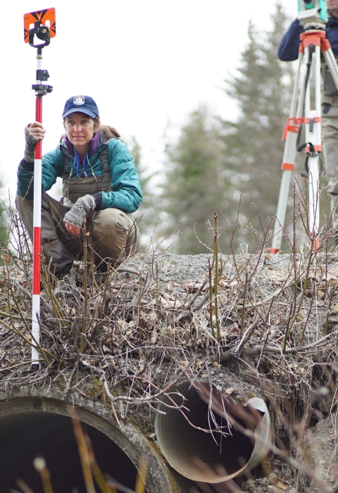 woman with survey gear squatting above a perched culvert