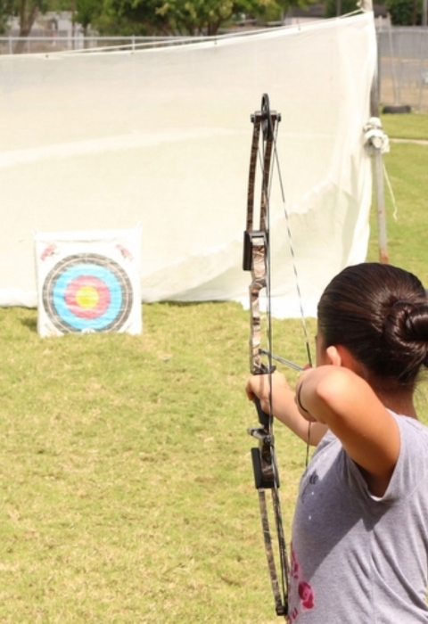 A young woman aims a bow and arrow at a target on a sunny afternoon.