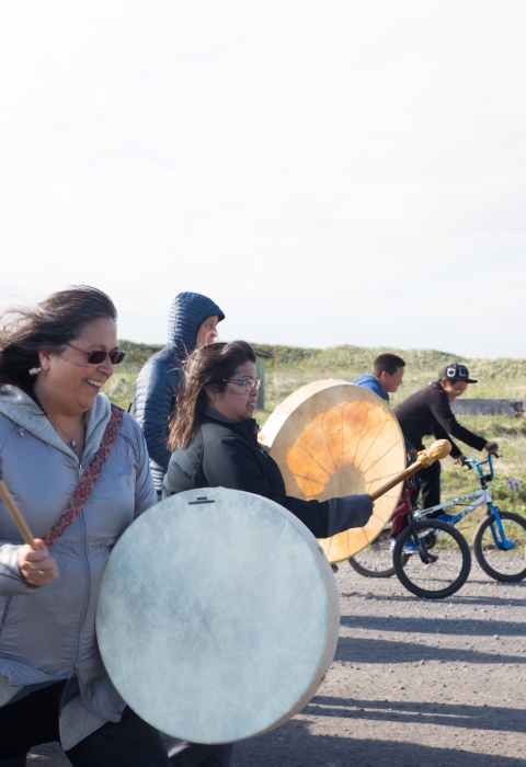 Woman with white  skin drum leads group of people walking down a road. Kids on bikes flank her.