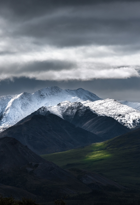 Snow-capped mountains loom over a green valley, all under stomy clouds