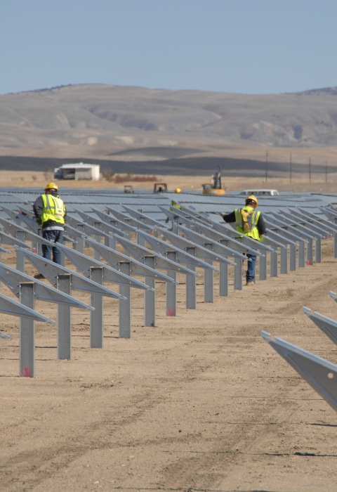 rows of gray solar panels stand out against the dry desert background. Solar panel installers wearing yellow vests work on the panels. 