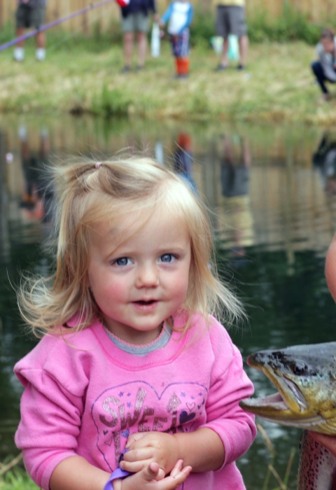 A very young blond-haired girl and an older girl pose with large trout in front of a pond where other children are fishing.