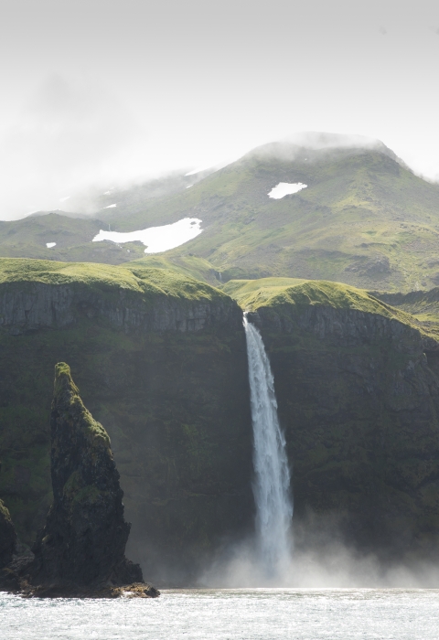 Waterfall plunges into the Bering Sea over a high cliff with foggy, snowy, and green mountain in the background.