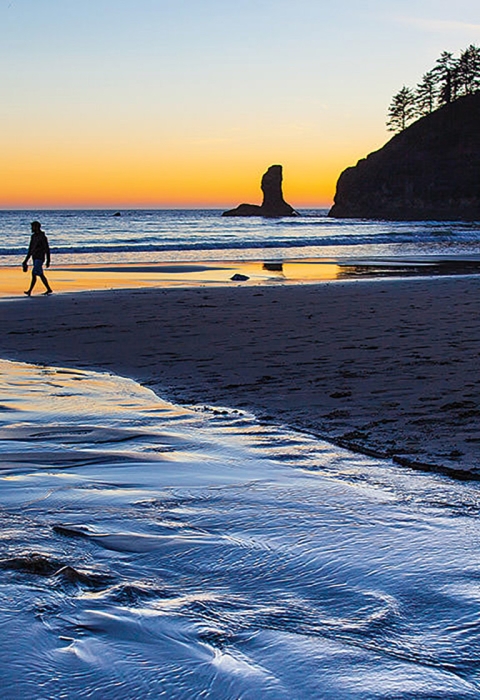 A stream flow down a beach to the ocean with silhouettes of humans walking and a distant sea stack at sunset